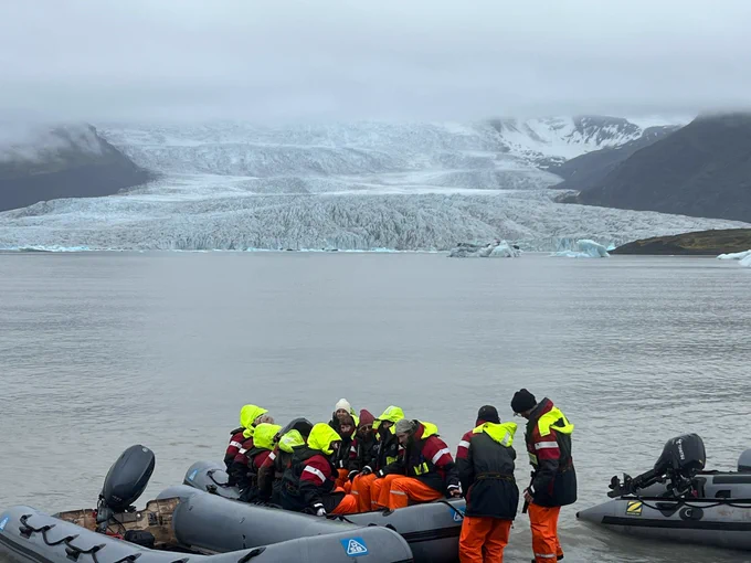 A group of tourists in safety gear gathers on a boat in front of a large glacier in a serene water body, with misty mountains in the background.