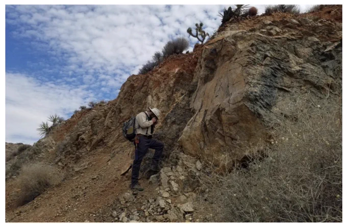 A man in field gear stands on a rocky slope near a cliff of exposed rock. He is looking closely a small piece of rock in his hand.