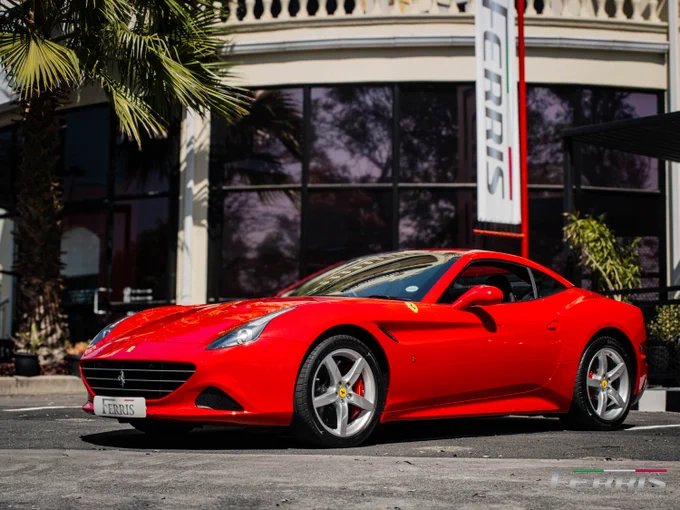 Red Ferrari California parked in front of a building with palm trees, under a clear sky. A Ferrari flag is visible in the background.