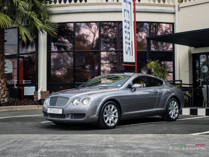 Silver Bentley Continental GT parked in front of a building with large glass windows and a palm tree nearby.