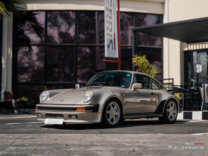 A classic silver Porsche 930 Turbo parked in front of a modern building with a Ferris banner, showcasing its sleek design.