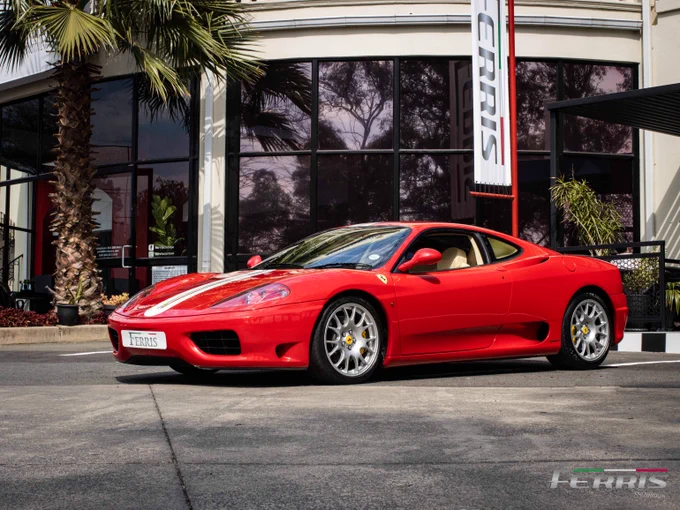 A red Ferrari sports car parked in front of a showroom with palm trees. The model is a Ferrari 360 with sleek design.