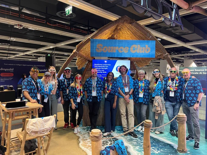 Group of people wearing matching blue floral shirts and colorful leis standing under a wooden hut sign labeled 'Source Club' at an indoor event.