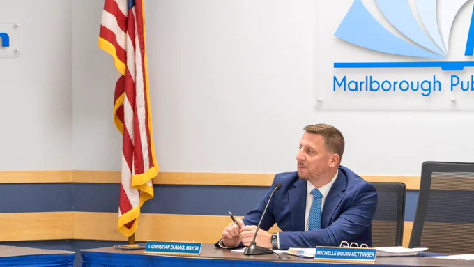 Marlborough School Committee Chair, Mayor Christian, sits at a table with the USA flag to his right and the MPS logo behind him