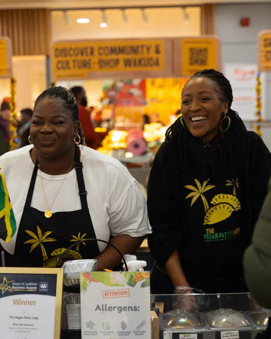 Two individuals at a market stall labeled "The Vegan Patty Lady," smiling and wearing tops with sunflower logos. A sign reading "Discover Community & Culture, Shop Wales!" is visible in the background.