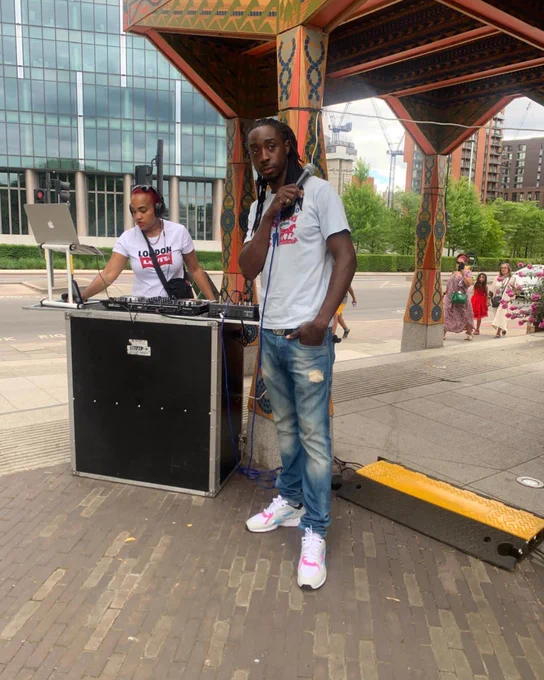 Two DJs at an outdoor event, one standing in front facing the camera and another focused on equipment. They're wearing matching 'I Love Soca' t-shirts.