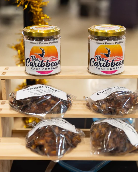 Products from The Caribbean Cake Company, including jars of Sweet Potato Pudding and bags of rock cake, displayed on a store shelf.