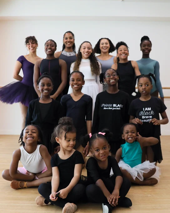 Group of diverse ballet dancers of different ages smiling in a dance studio, some wearing shirts with empowering messages.