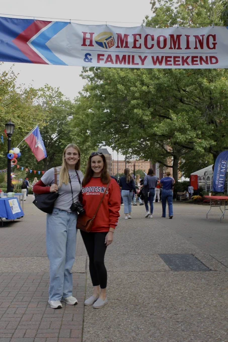 Two female students are standing in front of a banner that reads "Homecoming and Family Weekend." They are both wearing SoE merchandise.