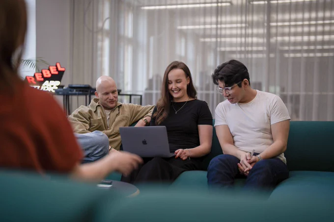 Three people are sitting on a green sofa in a modern office, looking at a laptop and smiling. A neon “Rebel App Studio” sign glows in the background. The atmosphere is casual and collaborative.
