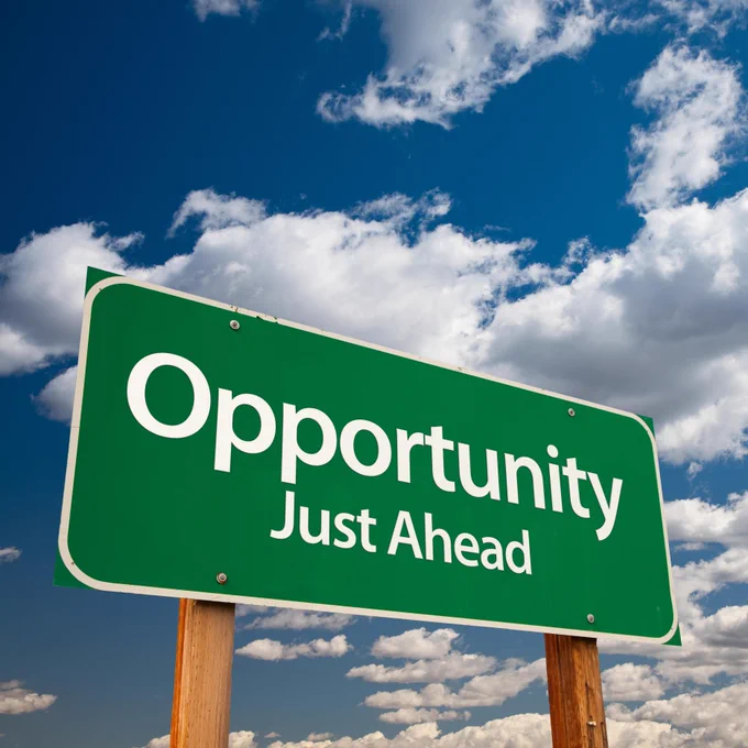 In front of a blue sky with white clouds, a green highway sign says 'Opportunity Just Ahead' in big white letters.