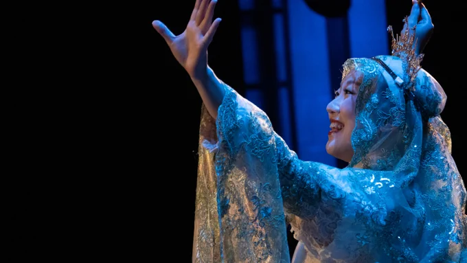 A performer in elaborate blue traditional attire and headpiece is gesturing expressively on stage.