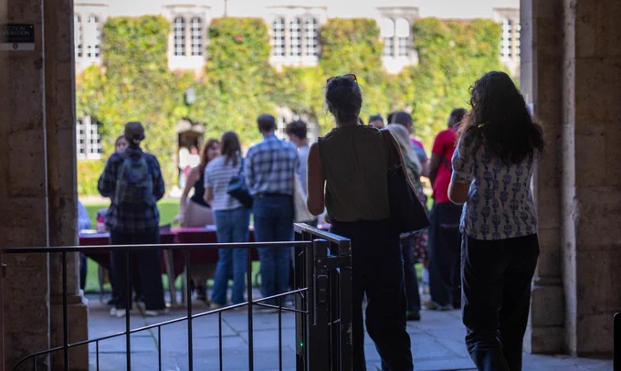 From the perspective of an entranceway, several people are seen walking out into the sunlit Front Quad of Exeter College. In the background, a historic, ivy-covered building provides a classic Oxford backdrop for a group of Open Day visitors.