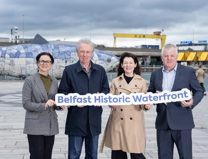 Four people stand in front of the Big Fish sculpture next to the River Lagan. They are holding a sign with text in blue Belfast Historic Waterfront in the distance is the Queens Quay Kiosk and Harland and Wolff cranes.