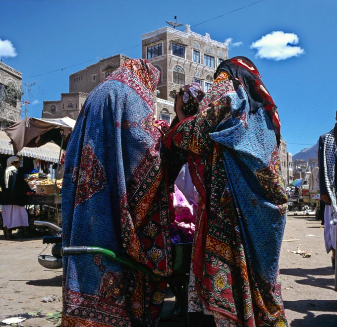 Two women wearing colourful patterned shawls stand closely together at an outdoor market, with traditional buildings and blue sky in the background.