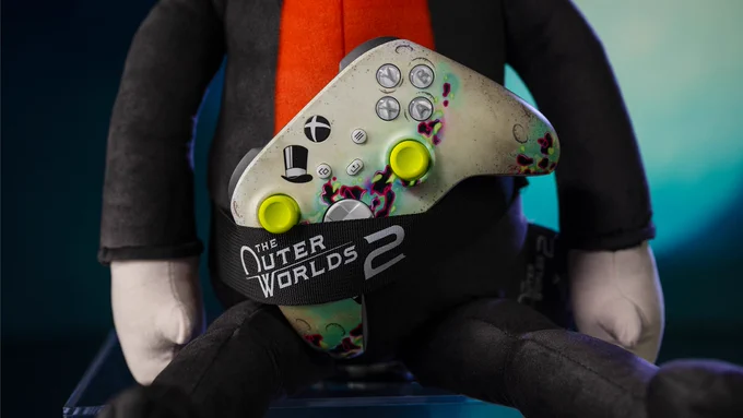 Close up image of the controller/remote slot holder that is a feature of The Outer Worlds 2 Moon Man Couch-Panion plush toy. In this image, the slot is holding an Outer Worlds 2 themed Xbox controller. The strap that holds the controller has the Outer Worlds 2 logo on it.