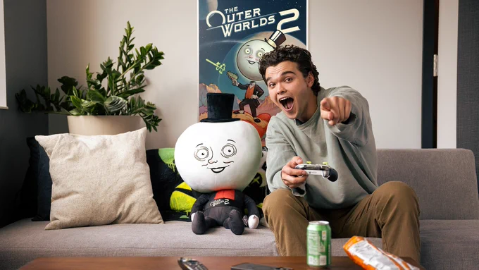 Image of a person sitting on a couch playing video games, pointing toward the TV with a smile and holding an Xbox controller. Also on the couch is The Outer Worlds 2 Moon Man Couch-Panion plush toy. Behind them is a poster for The Outer Worlds 2. On the table in front are a remote, a phone, a soda can, and an open bag of chips.