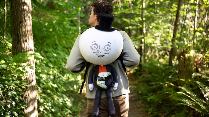 Image of a person walking through a forest trail surrounded by trees. The person's back is facing the camera and they are using The Outer Worlds 2 Moon Man Couch-Panion plush toy as a backpack. An Xbox controller is being held by the plush toy.