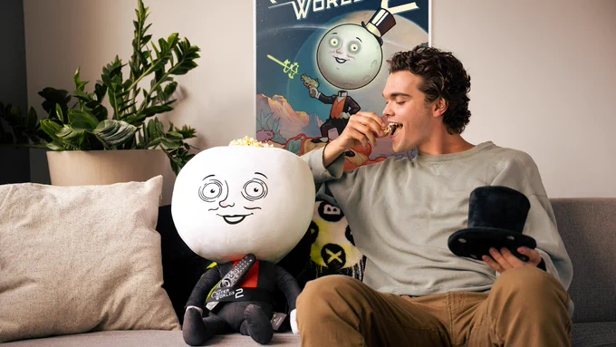 Image of a person sitting on a couch next to The Outer Worlds 2 Moon Man Couch-Panion plush toy. The person is holding the plush toy's top hat while eating popcorn out of the plush toy's popcorn bucket on top of its head. Behind them is a poster for The Outer Worlds 2. The TV remote is being held by the plush toy