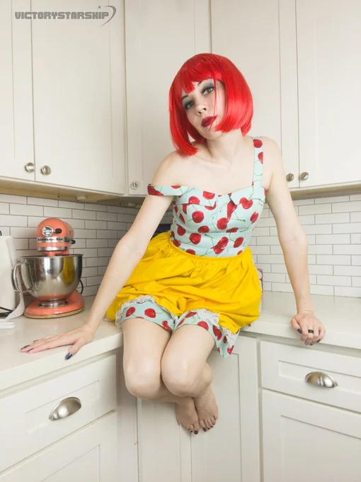 Cute girl doing a self portrait photoshoot. Standing in the kitchen, or sitting on the counter, with a stand mixer behind her. She is wearing a bright red wig, a blue dress covered in big red cherries, and a yellow apron. Retro cute fun whimsy.