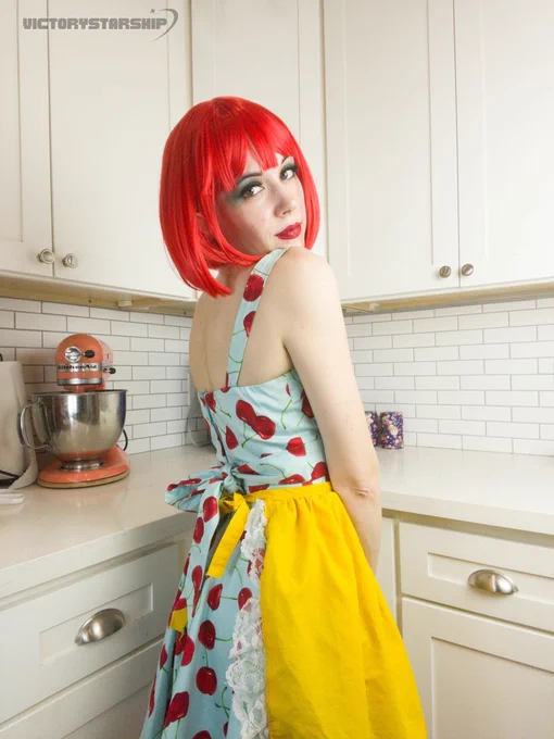 Cute girl doing a self portrait photoshoot. Standing in the kitchen, or sitting on the counter, with a stand mixer behind her. She is wearing a bright red wig, a blue dress covered in big red cherries, and a yellow apron. Retro cute fun whimsy.