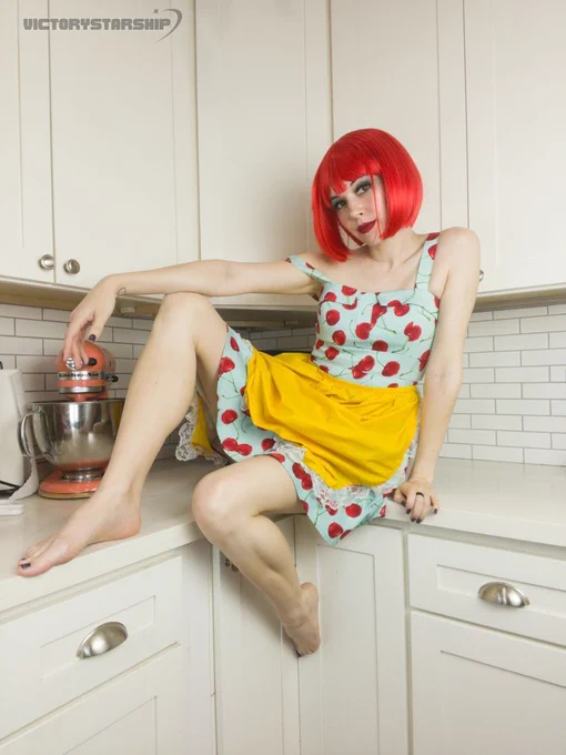 Cute girl doing a self portrait photoshoot. Standing in the kitchen, or sitting on the counter, with a stand mixer behind her. She is wearing a bright red wig, a blue dress covered in big red cherries, and a yellow apron. Retro cute fun whimsy. Barefoot with her feet on the counter.