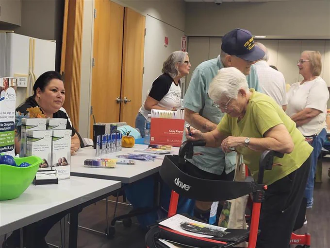 An older lady is placing some items in her bag and standing in front of a booth with her walker at the Southwest Florida Council of the Blind White Cane Safety Day event. 