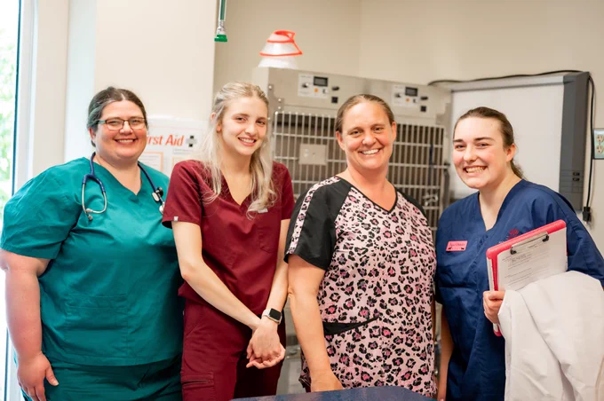 Four veterinary staff members smiling in a clinic, standing in front of kennels, wearing medical scrubs.