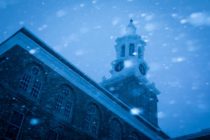 A low-angle shot captures a brick building with a white clock tower under a heavy snowfall. The clock face is visible, and snow is prominent against the blue-toned sky.