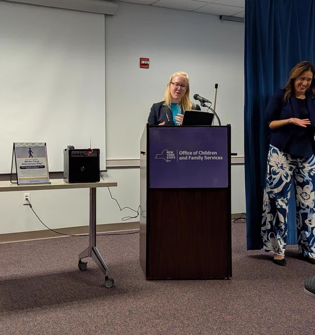 A photo of Kathyrn Carroll, a white woman with blonde hair wearing glasses and a blazer, speaking at a podium with a sign in front that says "Office of Children and Family Services". To her right is a sign language interpreter wearing a blue blazer and floral pants, smiling. On a desk to Kathryn's left is a sign that says "White Cane Awareness Day!"