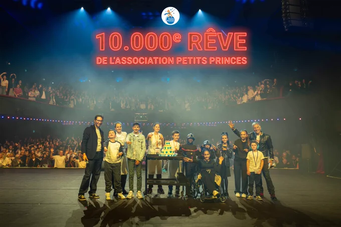 La photo montre une scène sur laquelle se trouvent Manu Levy et Nagui accompagnés de plusieurs enfants de de l'Association Petits Princes. Ils se trouvent sur la scène de l'Olympia. Derrière eux, les 1200 invités sont debout et lèvent les bras en l'air. Au-dessus d'eux apparaît un néon rouge où il est écrit "10000eme rêve de l'association Petits Princes". Enfin, on voit tout en haut de l'image le logo de l'association Petits Princes.