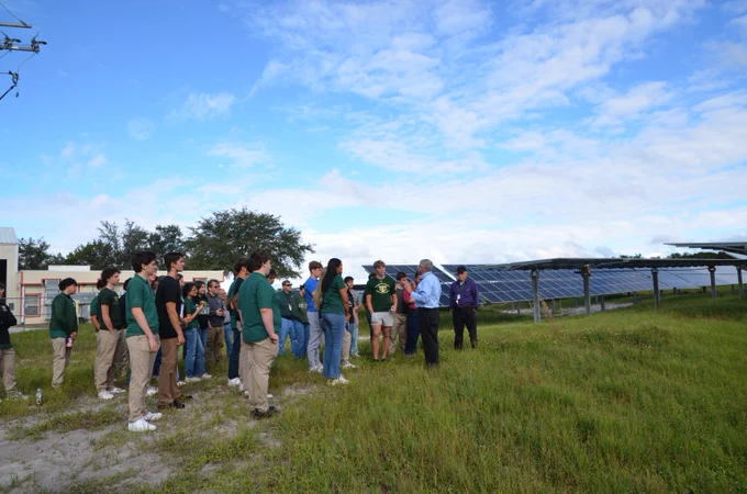 A group of high school students stands in a solar facility with an adult male who is speaking to them. They are on grass and in front of solar panels.