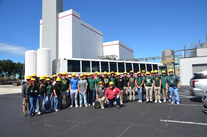 A group of high school students wearing hard hats poses in front of their bus at a power plant.