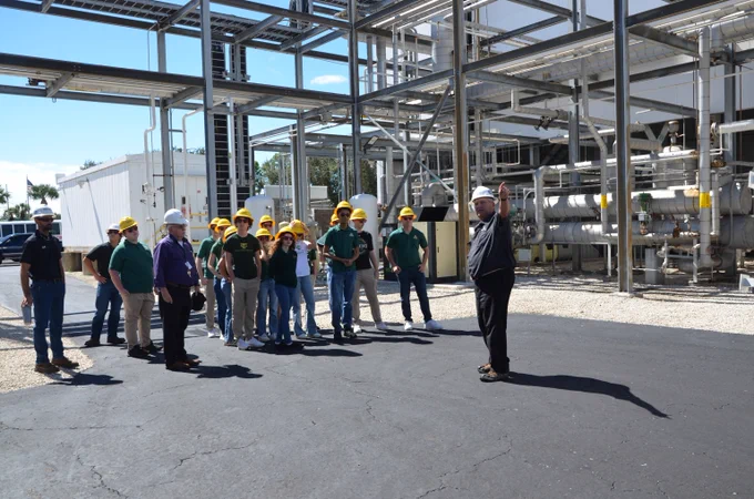A group of high school students wearing hard hats is outside while touring a power plant. They are listening to a man as he presents. He is standing in front of the group and gesturing with his arms as he explains the facility.