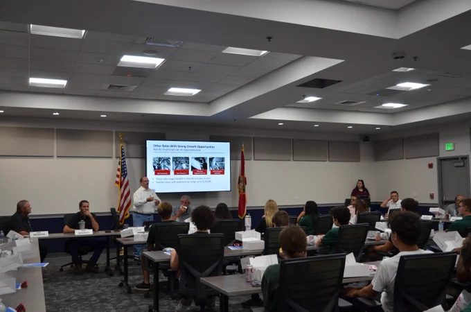 A man stands at the front of a board room to present to high school students. He is in front of a screen that shows a Power Point slide about careers in energy that may be available after completing vocational training or an apprenticeship.