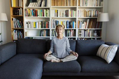 A picture of a woman sitting on a sofa with her eyes closed in the lotus position with a bookcase behind her
