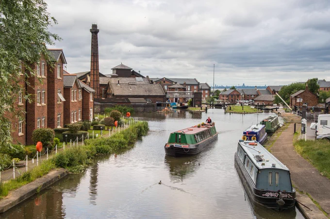 Canal boat travelling through the water at Ellesmere Port, surrounded by moored boats, red-brick buildings, and the tall chimney of the old pumphouse in the background.