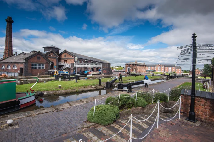 Volunteer helping a boater open a lock gate at the National Waterways Museum in Ellesmere Port, with the pumphouse and historic brick buildings in the background.