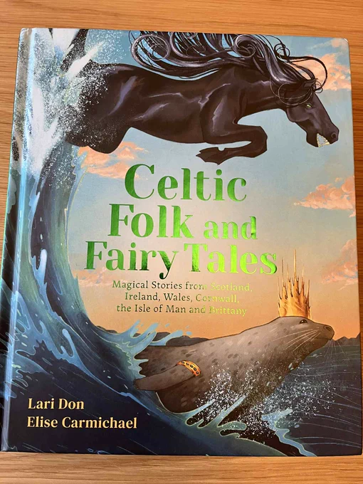 A hardback book laid flat on a wooden table. The title of the book, in shiny green foil in the centre of the cover, is Celtic Folk and Fairy Tales, and two names at the bottom left are: Lari Don and Elise Carmichael. The cover art is a black horse with wild mane and sharp teeth leaping across the top, a seal wearing a pointy gold crown swimming at the bottom, and a wave of blue and white curving up the left side of the cover, linking them.  There is a subtitle in smaller font reading: Magical Stories from Scotland, Ireland, Wales, Cornwall, the Isle of Man and Brittany.