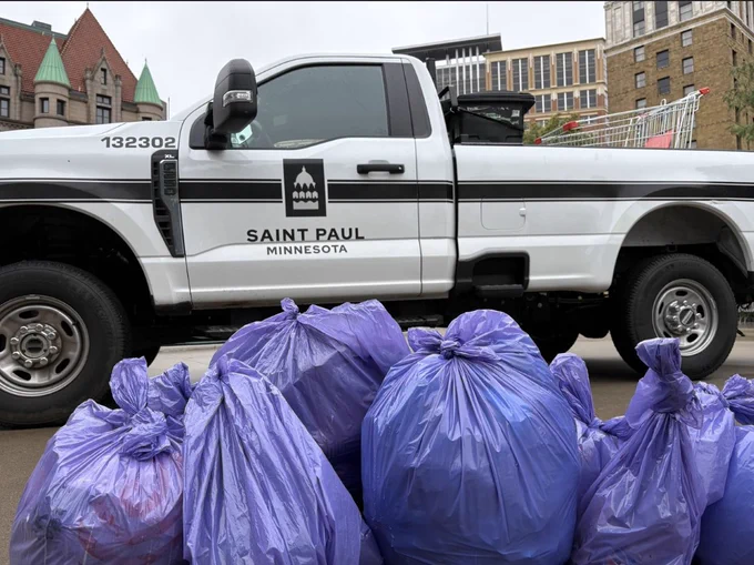 A white Saint Paul, Minnesota municipal truck is parked on a city street. In the foreground, several large purple trash bags are piled on the sidewalk. A shopping cart is visible in the truck's bed. Historic and modern buildings are in the background.