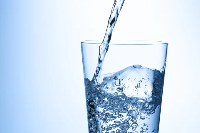 Clear water being poured into a transparent glass filled with ice cubes against a light blue background.