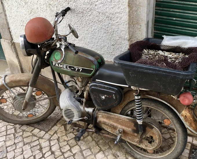 Ein ziemlich verrostetes Moped mit einem alten roten Helm auf der Lenkstange und einer schwarzen Plastikschale auf dem Gepäckträger. In der Schale liegt ein Netz und darin befinden sich Muscheln, kleine graue. Auf dem grünen Tank des Mopeds steht Famel-77 als Typenbezeichnung. Das Moped steht auf Kopfsteinpflaster vor einer weißen Hauswand.