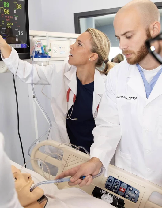 Two nursing students in a medical setting stand over a mannequin in a hospital bed.