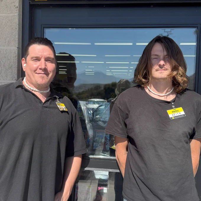 Two employees stand outside the store. One wears a polo and has short hair and a white shell necklace. The other employee has long hair, wearing a shirt.