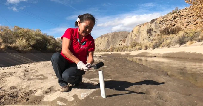 Karletta Chief, in a red shirt and gloves kneeling by a riverbed, using a tool to conduct hydrological research, surrounded by a scenic, rocky landscape under a clear sky.