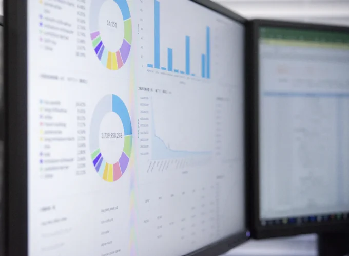 Photo of two computer screens showing data in pie charts, graphs and tables