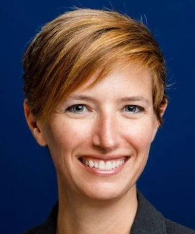 A smiling woman with short brown hair and blue eyes against a blue wall