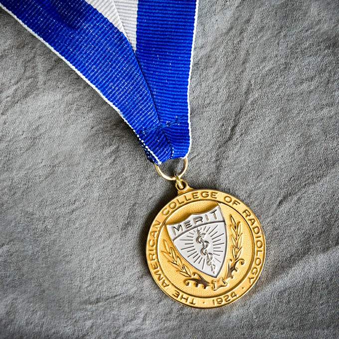 Gold merit medal from the American College of Radiology with a blue and white ribbon, lying on a grey fabric.