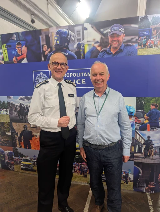 The image is of the Metropolitan Police Commissioner, Sir Mark Rowley standing next to Councillor Barry Lewis while they were at Sutton's Community Crime Fighting event. They are standing in front of a large sign which says Metropolitan Police with various images of police officers on it.