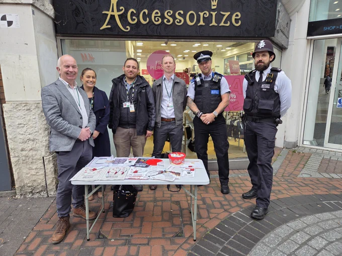 The image is of Leader of the Council, Cllr Barry Lewis, two members of staff from Sutton council's community safety team, Deputy Leader of the Council, Cllr Barry Lewis and a Sutton PCSO and police officer standing behind a stall in Sutton High Street for hate crime awareness week. In front of them is a small table with leaflets and information about hate crime awareness week.